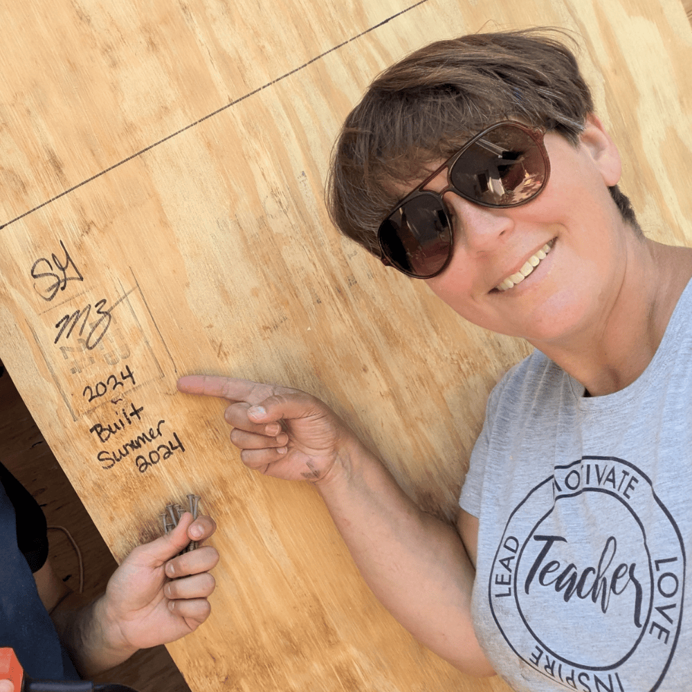 Picture of Sadie Gunnink wearing sunglasses and smiling while pointing at initials and the words, "Built Summer 2024" scrawled on the side of a wall.